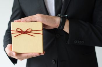 Close-up of man with wristwatch holding gift box while presenting new product. Unrecognizable businessman in black suit selling Christmas goods. Holiday concept